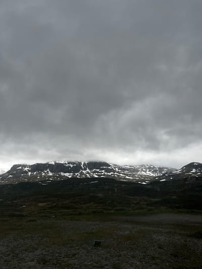 The image depicts a vast, mountainous landscape under a heavily overcast sky. The mountains, which are snow-capped, dominate the middle ground, their jagged peaks partially obscured by clouds. The foreground consists of a dark, rocky terrain with sparse vegetation. The overall impression is one of a wild, remote, and somber natural environment. The location is described as Unknown, Norway.