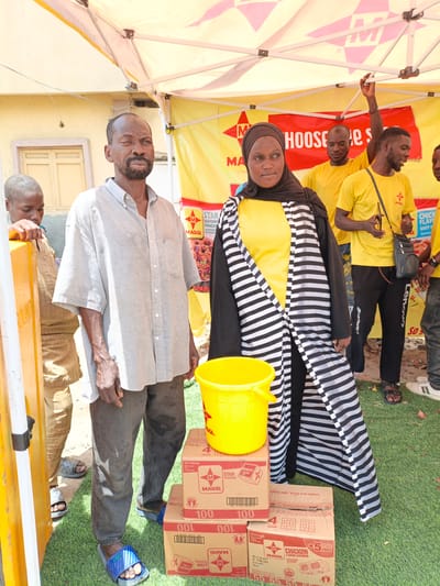 This image captures an outdoor scene, likely a promotional event or distribution in Tarauni, Nigeria, under a bright, sunny sky, suggested by the strong shadows and the tent providing shade. The setting features a yellow and white tent with branding, including the "MAGGI" logo prominently displayed. The ground is covered with artificial green turf, and several stacks of "MAGGI" seasoning boxes are visible, indicating a distribution or sale of these products.

In the foreground, a man with a beard and a light grey, short-sleeved button-down shirt stands to the left, facing slightly towards the camera with a neutral expression. Beside him, a woman wearing a black hijab and a yellow top, layered with a black and white striped open abaya, looks away from the camera. To their left, a younger person in a beige outfit leans on a yellow post. In the background, several other people in yellow t-shirts are visible under the tent, some appearing to engage with attendees or organize products. One man in the background raises his hand towards the tent.

A large yellow bucket sits on top of some of the stacked "MAGGI" boxes. The visible text on the boxes includes "MAGGI STAR SEASONING CUBE," "CHICKEN FLAVOUR SEASONING," and nutritional information such as "20x100x4g." The overall atmosphere appears to be one of community engagement or a commercial activity related to food products.