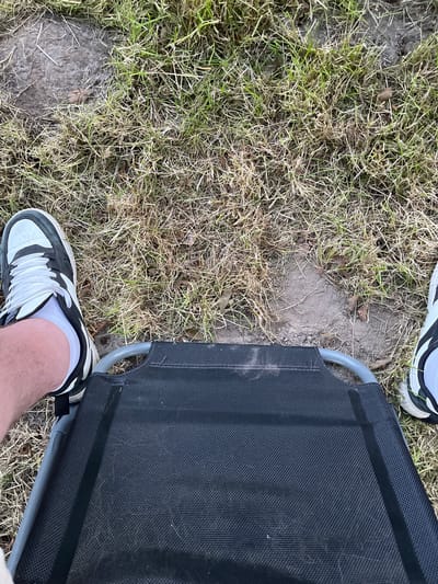 The image shows a first-person perspective of someone sitting on a portable chair, with their legs and feet visible. The chair is black and made of fabric, with grey metal supports. The person is wearing white socks and black and white sneakers. They are outdoors, with a background of dry grass and patches of bare earth. There are no identifying landmarks or features that would indicate a specific location in Uithuizen, Netherlands, or any other city. The scene appears to be a casual, everyday setting.