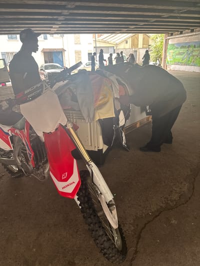 Underneath a concrete overpass in Saint-Denis, France, a person wearing a black baseball cap and a black t-shirt stands next to a white and red dirt bike. The dirt bike is facing left, with its front wheel towards the camera. Behind the dirt bike, a white pallet is piled high with clothing. To the right of the pallet, another person, wearing a dark striped sweater and black pants, is bent over, appearing to sort through the clothing. In the background, several other people are visible, and a mural depicting a landscape scene is painted on a wall to the far right. Buildings and trees can be seen beyond the overpass.
