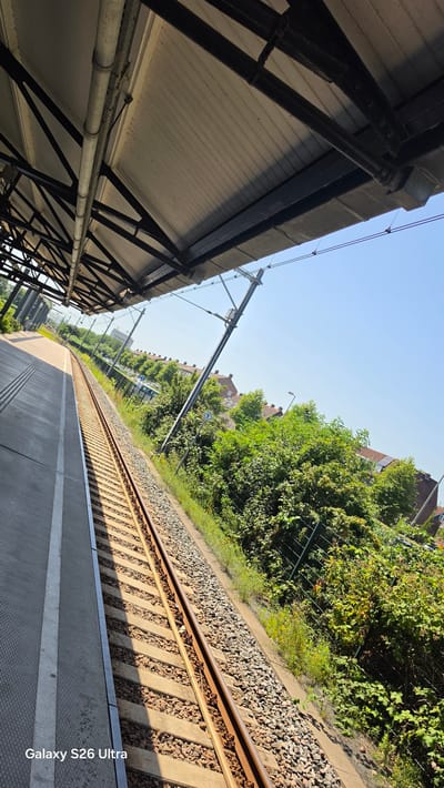 The image captures a view from a train platform in Vlaardingen, Netherlands. The perspective is from ground level, looking down a set of train tracks that curve into the distance. To the left of the tracks is a covered platform with a corrugated metal roof supported by dark metal beams. The roof extends overhead, creating a shaded area on the platform. The platform itself is made of concrete and has textured markings.
To the right of the tracks, lush green trees and bushes are densely packed, creating a natural barrier. Behind the foliage, several residential buildings with reddish-brown roofs are visible, suggesting an urban or suburban setting adjacent to the railway. The sky above is clear and bright blue, indicating a sunny day, likely in the late morning or early afternoon due to the distinct shadows cast by the platform and overhead structures.
Overhead, electrical wires for the train system are strung across the scene, supported by poles. A small blue and white sign with a symbol is visible on a pole amidst the greenery. The train tracks are laid on a bed of gravel and supported by concrete sleepers. The overall scene evokes a sense of an ordinary day at a functional public transportation hub, bathed in natural light. There is no discernible activity or people visible in the image. The watermark "Galaxy S26 Ultra" is present in the bottom right corner of the image.