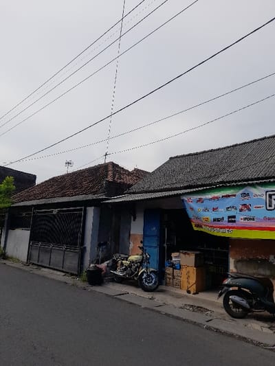 The image shows a street in Kecamatan Tanggulangin, Indonesia (latitude: -7.505, longitude: 112.716). The street is paved and runs from left to right in the image. There is a house on the right side of the street. The house has a tiled roof and a concrete facade. There is a large, black metal gate in front of the house. The house has a shop front with a blue door and windows. There is a sign above the shop front with the words "TDR" and "WIND" on it.  A yellow motorcycle is parked in front of the shop. A black scooter is parked in front of the shop on the right side of the image. There are several black power lines strung overhead across the image. The sky is overcast. There is no visible vegetation in the immediate foreground.
