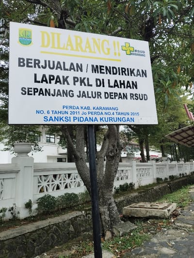 A sign in Indonesian, mounted on a black pole, is attached to a white concrete wall in Telukjambe, Indonesia (latitude: -6.321, longitude: 107.292). The sign is a warning prohibiting selling or establishing street food stalls on the grounds of the local hospital,  "RSUD Karawang". The text reads: "Dilarang!! (Forbidden!!) Berjualan/mendirikan lapak PKL di lahan sepanjang jalur depan RSUD Perda Kab. Karawang No. 6 Tahun 2011 Jo Perda No. 4 Tahun 2015 Sanksi Pidana Kurungan". It is a sunny day.  The sign is positioned in front of a tree, and there is a white concrete fence and a stone drainage ditch on the right. The sign and the fence are in front of a white building with windows.  The sign is in focus, while the background is blurred.