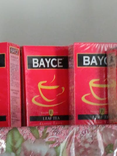 The image shows three red boxes of "BAYCE" brand tea arranged side-by-side on a surface with a floral pattern. The boxes are wrapped in clear plastic. The front of each box prominently features the word "BAYCE" in white text within a black rectangle. Below the brand name, there is a stylized yellow drawing of a teacup with steam. Some boxes also have additional text such as "LEAF TEA" and "CLASSIC TASTE." The lighting suggests an indoor setting, with no indication of time of day or weather. The overall impression is of a retail display or personal collection of tea products. The location context provided is Razzakov, Kyrgyzstan.