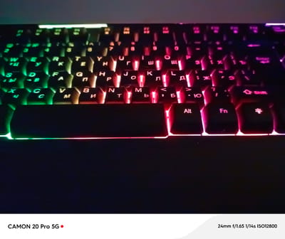 The image shows a close-up, low-angle view of a backlit keyboard with Cyrillic characters. The keys are illuminated by vibrant, multi-colored LED lights, ranging from green on the left to red on the right. The focus is on the keys, with some blurriness in the background. The bottom of the image displays text indicating "CAMON 20 Pro 5G" with a red dot, and camera settings "24mm f/1.65 1/14s ISO12800". There is no visible indication of the city of Yakutsk, Russia, or any people, actions, or interactions within the frame. The environment appears to be indoors, likely a desk setup.