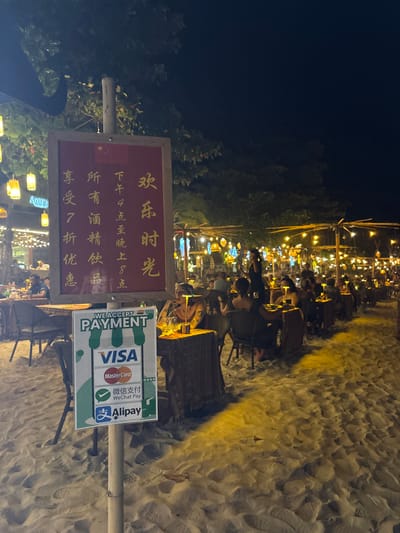 This image depicts an illuminated outdoor dining establishment situated on a sandy beach at night in Ban Ko Li Pe, Thailand.
In the foreground, a light-colored wooden pole supports two signs. The upper sign is a large, red rectangular board with gold Chinese characters, indicating "Happy Hour" from 4 PM to 8 PM with a 70% discount on all alcoholic beverages, and features a small Chinese flag at its top. Directly below it, a smaller vertical sign with a white and green background states "WE ACCEPT PAYMENT" and displays logos for VISA, MasterCard, WeChat Pay, and Alipay.
The dining area extends into the midground and background, comprising numerous small, square tables arranged in rows directly on the sand. Each table is covered with a patterned fabric and features a small, illuminated portable lamp. Dark chairs are positioned around these tables. Overhead, an extensive network of string lights and various decorative lanterns, emitting warm yellow and orange light, illuminates the entire scene.
Multiple individuals, approximately 20 to 30, are seated at the tables, engaged in dining or social interaction. One figure, possibly staff, is observed standing in a dark uniform near the center-right, appearing to attend to guests. Dark tree foliage is visible against the deep black night sky, bordering the illuminated dining area. The ground surface consists entirely of light-colored sand.