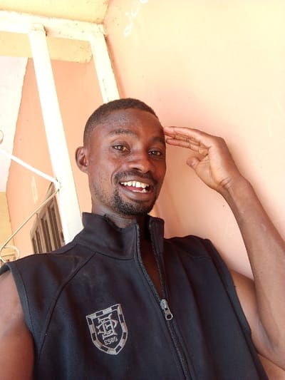 A dark-complexioned adult male is depicted in a close-up portrait within the city of Otokiti, Nigeria. He wears a black sleeveless zippered vest with a grey and white shield-shaped embroidered emblem on the left chest, featuring intertwined "SD" letters above "ESGO" text. The man has short, dark hair and a short beard, and is smiling broadly, exposing his teeth. His right hand is raised to his forehead. The background consists of a light orange-peach colored wall with faint white markings on its upper right portion. Partially visible on the left are white or light-colored metal railings and a dark window or opening.