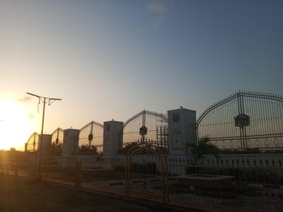 A low-angle, medium shot depicts a series of ornate, arched gates stretching into the distance under a sky gradient from yellow-orange near the horizon to pale blue overhead. The gates are constructed from dark metal, with a decorative flourish at the top of each. They are interspersed with squat, cream-colored pillars adorned with golden-yellow embellishments. Behind the gates, green foliage is visible. A lower fence of golden-yellow metal runs along the foreground, obscuring the view of what appears to be a landscaped area. To the left, a utility pole rises against the bright sun. Scaffolding is visible behind one of the pillars, suggesting ongoing construction or maintenance. The scene is washed with warm light, indicating sunrise or sunset. Based on the prompt, the location is Tawpo, Nigeria.