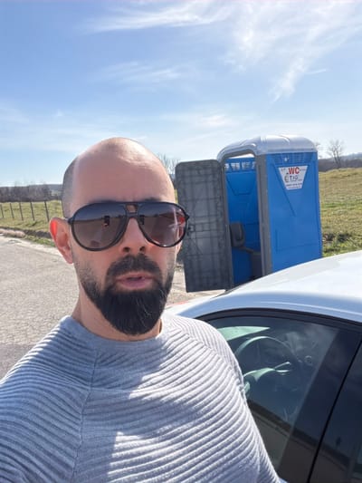 A front-facing image captures a male subject taking a selfie outdoors in Dositeevo, Bulgaria. The man, appearing bald with a dark, full beard and wearing dark aviator-style sunglasses, is dressed in a light grey horizontally ribbed long-sleeved sweater. He stands on an asphalt surface, positioned to the left of a white vehicle, likely a sedan, with its driver's side visible. Behind the vehicle and the man, within a grassy field, a blue and grey portable toilet with an open door is present. The portable toilet bears the inscription "WC T&G" and the phone number "0887 014 416" on its side. A wire fence separates the asphalt surface from the grassy field, which extends to a distant treeline under a bright blue sky with sparse white clouds. The scene is brightly lit by natural sunlight.