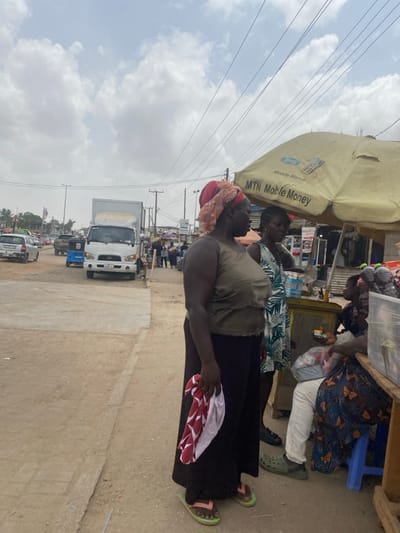 The image depicts an active street market scene in Ablekuma, Ghana, under an overcast sky. In the foreground, a dark-skinned woman with a medium build stands right of center, facing slightly right. She wears a light olive green sleeveless top, a dark long skirt or trousers, and light green flip-flops with pink straps. A reddish-orange and cream patterned headwrap covers her head. She holds a folded white cloth with red geometric patterns in her right hand.

To her immediate right and slightly behind her, another dark-skinned woman stands, wearing a green, white, and dark-patterned sleeveless dress. Further right, beneath a large yellow umbrella branded with "MTN Mobile Money," is a small wooden vendor stand. An individual, partially visible, is seated on a blue stool at the stand, wearing white trousers and a dark blue patterned garment, with dark green Crocs on their feet. A child with dark hair and a dark top is partially visible near the stand. The stand displays various goods, including multiple plastic bottles of dark liquid, a blue plastic water bottle, and packaged items in clear bags.

The ground in the immediate foreground is a mix of dirt and concrete. In the mid-ground, a paved road stretches diagonally from left to right, populated by several vehicles. A white box truck, identified as Hyundai, faces the camera in the center. Behind it, a blue motorized tricycle is visible. To the far left, a silver hatchback car moves away from the camera. Multiple utility poles and overhead power lines crisscross the sky. In the background, additional pedestrians and market stalls are visible, indicating a busy thoroughfare. The sky is largely covered with white and grey clouds.