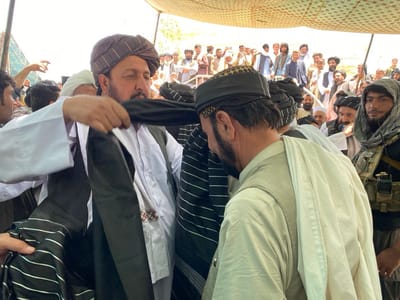 The image shows a group of men gathered under a tent, likely in a public space, at the location specified by the coordinates in the city of Zaker-E Sharif, Afghanistan. The scene is one of a gathering or a meeting, with several men standing and talking. The men are dressed in traditional Afghan clothing, with many wearing turbans. One man is wearing a military-style vest with a radio attached. The man at the center of the image is wearing a black turban and a white robe, and is placing a black scarf or cloth over another man's shoulders, who is wearing a white shirt and a traditional hat. The men are surrounded by a crowd, some of whom are standing and some of whom are sitting. The background shows a large group of people, seemingly waiting or watching.  The environment is open and appears to be a public space or square. The mood seems to be one of seriousness and respect.