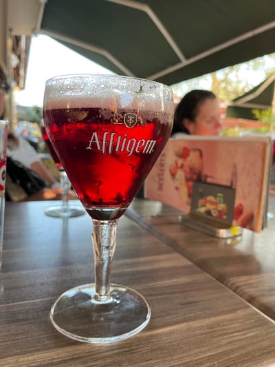 A red Affligem beer in a stemmed glass sits on a wooden table at an outdoor cafe. A person is visible behind the glass, near a menu stand. The cafe appears to be located in Montélimar, France, based on contextual clues. The surrounding environment suggests an outdoor setting with partial shade from an awning.