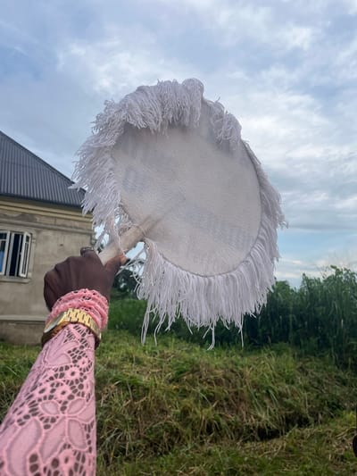 A vertically oriented outdoor photograph captures a dark-skinned hand holding a round, white hand fan against a natural backdrop in Aghalokpe, Nigeria. The fan, serving as the central subject, features a solid white circular surface with long, white, thick fringe details along its entire circumference. The hand grips a light-colored, possibly wooden, handle at the bottom of the fan.

The person's arm, adorned with a light pink long-sleeved garment, extends from the right side of the frame. The sleeve is made of a fabric with an intricate lace or cut-out floral pattern, revealing hints of skin underneath. On the wrist, a gold-toned watch with a classic metal link band is visible, alongside multiple stacked pink bracelets, possibly made of beads or crochet.

The background presents a cloudy sky, suggesting it is daytime, possibly overcast or late afternoon. Below the sky, lush green grass and dense green foliage are visible, indicating an outdoor, natural environment. To the left, parts of a building can be seen, featuring a dark grey sloped roof and light-colored walls with a window. The overall setting implies a scene from a rural or suburban area in Nigeria. No discernible text is visible in the image.