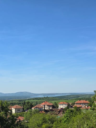 The image shows a view of Bokilovtsi, Bulgaria. The right side of the image depicts houses with red tile roofs nestled amidst lush green trees. In the distance, a river flows through a valley with a mountain visible in the background. The left side of the image is dominated by a clear blue sky.