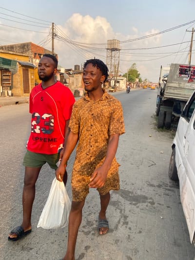 Two men are standing outside on an asphalt road. One man wears a red shirt, while the other wears a tan patterned button-up shirt. Both are smiling. They are holding a white plastic shopping bag. Behind them is a busy street with a large truck. A utility pole and water tower are also visible in the background.