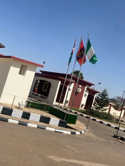 The scene depicts the exterior of a facility in Tudun Nupawa, Nigeria, under a clear blue sky on a sunny day. In the midground, three flags on silver poles are unfurled: from left to right, a green, white, and red flag with an emblem (likely Kaduna State), a predominantly red flag with a golden emblem (likely Nigerian Army), and the Nigerian national flag (green, white, green). Behind the flags, multiple one-story buildings with white walls and dark red trim/roofs are visible, featuring dark windows and an exterior air conditioning unit on one structure. A paved area with light-colored pavers and an artificial green turf section sits in front of the buildings. A black and white checkered curb separates this area from an asphalt road in the foreground, which bears a white arrow pointing left. Several palm trees are visible in the background, along with a conical evergreen tree to the right of the flags. No individuals are present.