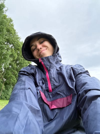 A young woman in a dark blue hooded rain jacket with a pink zipper and accent panel is looking up at the camera. She has dark hair and is smiling. The jacket appears wet, with water droplets visible on the fabric. The background shows a lush green tree on the left and a pale, overcast sky occupying the upper portion of the image. A lamppost is partially visible behind the tree. The foreground is dominated by the woman's jacket.