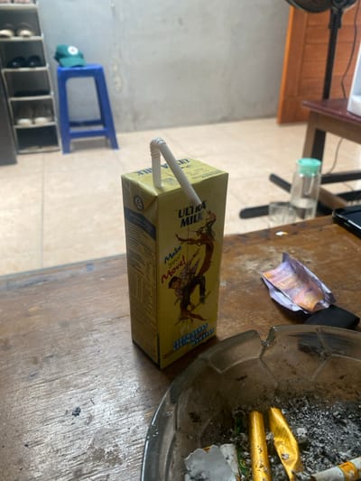 A yellow Ultra Milk carton, with a white flexible straw inserted into its top, rests on a worn, light-brown wooden table. The carton features graphics of a person on a skateboard and the text "Make Your Move!" To the right of the carton, a dark, circular ashtray, patterned with a soccer ball design along its rim, contains ash, several cigarette butts, and two crumpled gold-colored foil wrappers. Adjacent to the ashtray, a crumpled Indonesian Rupiah banknote and a small dark object are visible.
The background shows an indoor environment with light-colored tiled flooring and plain walls. On the left, a multi-tiered shoe rack holds several pairs of shoes. Behind it, a blue plastic stool supports a green baseball cap. On the right, a portion of a dark wooden table is visible, holding a clear bottle with a light green cap, a clear glass, and the base of an electronic fan. A closed wooden door is partially visible in the far right background. No individuals are present. The scene is located in Cimenyan, Indonesia.