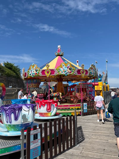This image shows a carousel on Llandudno Pier in the United Kingdom. The carousel is decorated with colorful patterns and cartoon characters and is surrounded by a wooden platform. People are standing on the platform, some of them are on the carousel, and there is a sign indicating that tickets are required to ride. The sky is blue with a few clouds. The sign says, "3 tokens each person" and "no loose or baggy clothing."