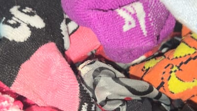 The scene depicts a close-up view of multiple tightly packed, crumpled textile items, consistent with an assortment of socks, located within the city of Lompoc, United States. No individuals are present, and no actions or interactions are depicted. The foreground and midground feature a pink ribbed fabric object. To the left, a black fabric item with distinct white stylized skull or bone patterns is visible. Above the pink fabric, a purple textile item, identified as a sock, displays a white Nike logo comprising the "NIKE" text and its characteristic swoosh design. Towards the bottom center, a grey fabric item with black and white striped or abstract patterns is visible. In the bottom right quadrant, an orange textile item features yellow and black patterns, possibly depicting cartoon characters or abstract designs. Various other dark and brightly colored fabric fragments are interspersed throughout the pile, indicating a mixed collection of apparel. The overall environment is an indistinct confined space, likely a drawer, hamper, or container.