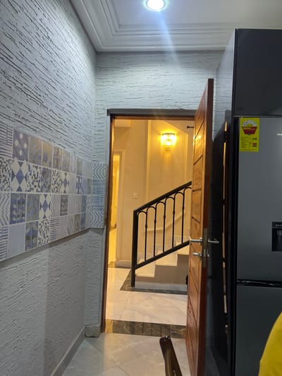 This interior photograph captures a modern living space, likely within a residence in Adenta Municipality, Ghana, illuminated by artificial lighting. The image is framed to show an open wooden doorway in the center, looking into a descending staircase. The staircase features light-colored treads and risers, complemented by a black metal balustrade with elegant scrollwork. A decorative wall sconce with a warm glow lights the stairwell.
To the right of the doorway, a large, dark-colored Hisense refrigerator is partially visible. It has a sleek, glossy finish and prominently displays a yellow "ENERGY GUIDE" label. Visible text on this label includes the brand "Hisense," the number "248" (likely indicating energy consumption), and an "H" logo. A sliver of a yellow-green cushion or piece of furniture can be seen to the far right, adjacent to the refrigerator.
The foreground wall, positioned to the left of the doorway, is finished with a textured, light-colored plaster. A significant portion of this wall is adorned with an arrangement of square decorative tiles. These tiles are rendered in shades of blue, white, and gray, showcasing a variety of geometric and abstract patterns. Overhead, a bright, circular recessed light fixture is visible on the white ceiling, which also features decorative molding. The floor leading up to and around the staircase is covered in polished tiles with a white and dark gray marble-like pattern. No people are present, and the scene is static.
