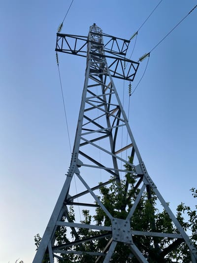 An outdoor, low-angle, medium shot captures a tall, metal power transmission tower against a clear, pale blue sky. The tower is constructed from a lattice of grey metal beams, bolted together with visible fasteners. At the top, the tower branches out to support thick, dark electrical wires. Clear, glass insulators are attached to the tower's arms, suspending the wires. Lush green foliage from trees peeks out from behind and below the tower, indicating a natural or semi-natural environment. The image provides no information about the presence of people or specific actions. The setting appears to be within or near the city of Grigoriopol, Moldova, given the provided context.