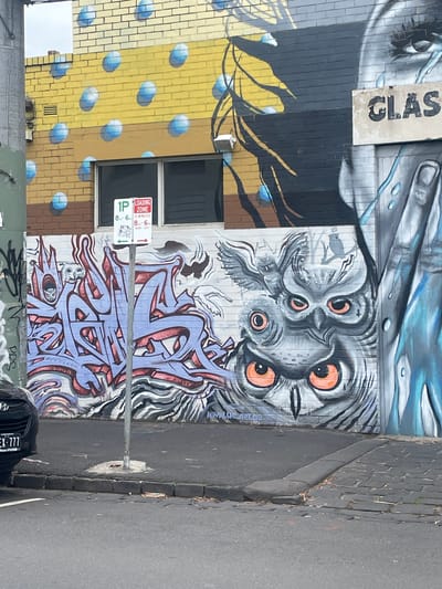 The image shows a street scene in Melbourne, Australia, dominated by a large, vibrant mural painted on a brick building. The mural features a stylized owl with large orange eyes in the center, flanked by graffiti-style lettering in purple and red on the left. To the right of the owl, the mural continues with what appears to be a large, abstract depiction of a human face with blue tears and a hand. The building itself is made of yellow and grey brick, with blue circular patterns painted on the upper section. A white sign with red lettering, indicating a "1P LOADING ZONE" with specific time restrictions, is mounted on a pole in front of the mural. A portion of a dark-colored car is visible on the left side of the image, parked on the street. The street is paved with asphalt, with a cobblestone section visible at the curb. The sky appears overcast.