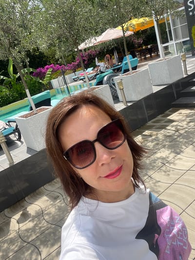 In this selfie, a woman with brown hair and sunglasses smiles while posing in an outdoor area with a swimming pool. She is wearing a white t-shirt with a graphic print on the sleeve. The background shows lounge chairs, trees, flowering bushes, and a building. The paving stones on the ground are patterned with shadows cast by trees. The scene appears to be a resort or hotel area, possibly in a warm climate given the presence of a pool and outdoor seating. The location is identified as Abaata, Abkhazia.