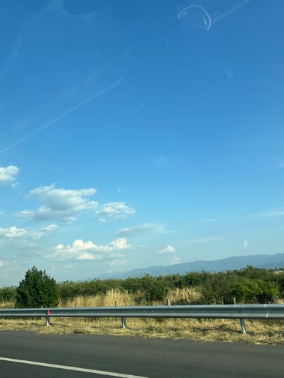 The media file depicts a roadside scene under a clear blue sky with scattered clouds. The perspective suggests the photograph was taken from inside a moving vehicle, likely looking out the side window. The right side of the image features a section of asphalt road with a white line marking the lane. Adjacent to the road is a metal guardrail, typical of highways or major roads. Beyond the guardrail, a strip of dry, yellowish grass and vegetation is visible, leading to a dense line of green trees and foliage that forms a backdrop. In the distance, a hazy mountain range can be discerned against the sky.
Several small, white clouds are dispersed throughout the sky, which is predominantly a bright blue. Some subtle streaks or smudges are visible on what appears to be the window through which the photo was taken, with one circular smudge prominent in the upper left quadrant. There are no people visible in the image. The scene appears to be set in a rural or semi-rural location, given the natural landscape and the absence of urban structures. The lighting suggests it is daytime, likely mid-morning to late afternoon, with the sun high in the sky. The overall impression is one of travel through a scenic, open landscape. No text is visible in the image.