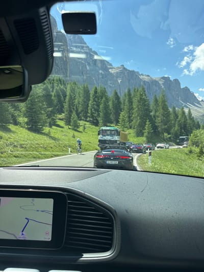 The image is taken from inside a vehicle, looking out through the windshield at a mountain landscape. The scene appears to be a winding road with a queue of vehicles. A cyclist is visible on the side of the road. The time of day is likely daytime, judging by the bright sunlight and clear blue sky with some clouds. The weather appears pleasant and sunny. The mountains are tall and rocky, covered in lush green pine trees, suggesting a mountainous region. The road is paved and marked with a white line.
In the foreground, the interior of the car is visible, including the dashboard and a navigation screen displaying a map. The navigation screen shows a route highlighted in blue, with a speed limit sign indicating 60. The license plate of the car closest to the viewer reads "K.HD318". Further up the road, there are several other cars, including a truck with "SCHMUTZIG" visible on its side.
The overall impression is a scenic drive through a mountainous area, possibly a popular tourist destination, with a bit of traffic congestion. The mood seems to be relaxed and appreciative of the natural beauty. The location cues like the license plate and the destination on the navigation screen suggest it might be in the Alps, specifically Val Gardena in Italy.