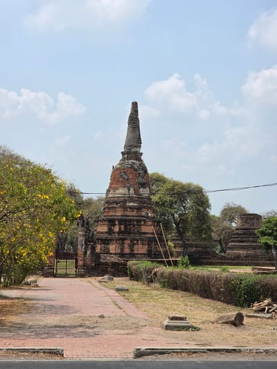 The image shows the ruins of a temple in Phra Nakhon Si Ayutthaya City Municipality, Thailand. The temple's main structure is a tall, brick chedi, partially crumbling, with a pointed spire. The lower sections of the chedi are surrounded by trees and other vegetation. A section of brick wall with a gate stands nearby. In the foreground, a tree with yellow flowers is partially visible. The background is composed of a blue sky with scattered white clouds. The foreground also includes a red brick walkway running parallel to the temple and wall, with an asphalt road further along. The overall scene depicts an ancient temple complex amidst modern surroundings.