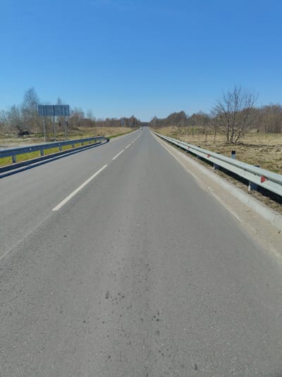 The image shows a straight, asphalt road under a clear, blue sky. The road is two-lane, with a solid white line separating the lanes. Metal guardrails are present on both sides of the road. The adjacent land is mostly bare, with some scrub vegetation and small trees. In the distance, a vehicle is visible on the road. The location is unspecified beyond being in Primorye, Russia.