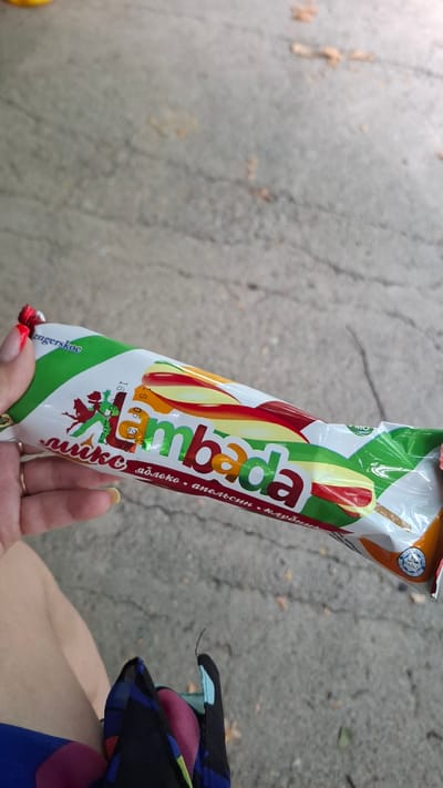 A person's hand with bright red nail polish is holding a wrapped ice cream bar. The ice cream bar has a white wrapper with green and colorful fruit imagery, and text that appears to be in Cyrillic script. The brand name "Lambada" is prominently displayed in large, multicolored letters on the wrapper. Below the brand name, there are words that likely describe the flavors. The background consists of a textured gray surface, resembling pavement or concrete, with some scattered organic debris. A small portion of the person's leg and a colorful scarf or fabric are visible in the lower part of the image.