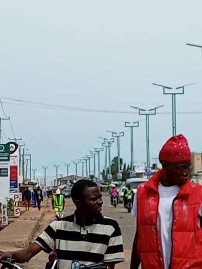 The image depicts a street scene in Garko, Nigeria, characterized by a row of modern streetlights lining both sides of the road. In the foreground, two men are prominent. The man on the right wears a red puffer vest over a white t-shirt and a red head covering adorned with white beads. The man on the left, partially obscured by the motorbike he is on, wears a black and white striped t-shirt. In the background, several other individuals are visible walking and riding motorcycles. One person in the middle ground wears a bright green vest, suggesting they might be a worker or security personnel. Vehicles, including cars and motorcycles, are present on the road. The environment appears to be an urban or semi-urban area with buildings and shops lining the street. The sky is overcast, suggesting a cloudy day. Signs, including one with "POS" visible, indicate commercial activity. The overall impression is of a bustling street in a Nigerian town.
