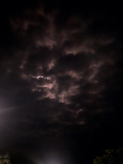 The image depicts a night sky filled with dark, textured clouds that appear to be illuminated from behind by a light source, possibly the moon or artificial lights. There are hints of pinkish hues within the clouds. In the lower left corner, a faint, bright light beam enters the frame from the left. The lower edges of the image show dark silhouettes of foliage, suggesting trees or other vegetation. The scene is dark and atmospheric, indicative of a nighttime setting. The location in Labissa, Indonesia, is a general context and not visually evident from the image itself.