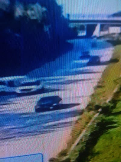 The image displays a heavily pixelated and low-resolution capture, purportedly depicting a scene in the city of Mista Ali, Nigeria. A prominent body of water, possibly a river or large canal, traverses the mid-ground horizontally. Adjacent to this water body is a light-colored surface, appearing to be a paved road or embankment. On this surface, in the lower-middle portion, a dark-colored vehicle, consistent with a car or small SUV, is present and casts a discernible shadow. To the left of this vehicle, several other lighter, indistinct shapes are visible on the same surface, possibly representing additional stationary objects or vehicles. In the upper mid-ground, a bridge-like structure with multiple supports spans across or is positioned above the water. The left side of the frame (upper portion) shows a deep blue area, suggesting a dense wooded area or a heavily shadowed land feature. The right side of the frame (lower portion) features an area with green and brown tones, indicative of terrain covered in vegetation, possibly a hillside or bank. The background (top left) is uniformly dark blue, suggesting an expanse of sky or distant land. No individuals, specific actions, or clear interactions are discernible due to the severe pixelation and lack of detail.
