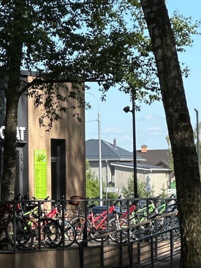 The image captures a row of parked bicycles in front of a building with a sign displaying "Лето" (Summer) in green text. The bicycles are positioned behind a metal railing, with some predominantly red and green frames visible. The building itself appears to be commercial, with large windows and a textured facade. Trees with lush green foliage partially obscure the view of the building and the sky. In the background, residential buildings with dark tiled roofs and chimneys are visible, suggesting a suburban or small town setting. Streetlights are also present in the background. The overall scene is bathed in natural daylight, casting shadows and highlighting the textures of the natural and man-made elements. The image is taken from a slightly elevated perspective, looking towards the building and bicycles.