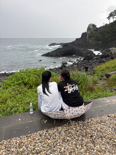 Two young women are seated on a stone path next to a rocky coastline, with lush green vegetation between them and the water. One woman, wearing a black t-shirt with a white logo, is crouched down, partially obscured by the other woman. The second woman, wearing a white t-shirt and blue shorts, is also crouched, facing away from the camera and towards the ocean. They are sharing an open umbrella with a pattern of oranges. A partially full water bottle rests on the path near the woman in the white shirt. The sea is a muted grey, with waves crashing against the dark rocks. The sky is overcast, suggesting a cloudy or rainy day. In the distance, a dramatic cliff face covered in dense green foliage rises from the sea, with what appears to be a small building or structure nestled on its summit. The ground is a mixture of dark, wet paving stones and a bed of small, multi-colored pebbles. The overall mood is one of quiet observation of the rugged coastal scenery. The visible text on the umbrella appears to be in a foreign language, possibly Korean, with the word "JEJU" faintly visible.