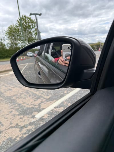 The image shows a side view mirror of a vehicle, reflecting the driver taking a photograph of the mirror with their own smartphone. Reflected in the mirror is the side of the vehicle to which it is attached, a line of traffic cones, a parked white car, an electrical pole and a tree. The road surface is visible outside of the vehicle, marked with a white line. The location is identified as Pity Me, United Kingdom.