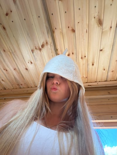 This image captures a close-up, angled selfie of a person with long, flowing blonde hair, partially covered by a white felt sauna hat. The person is making a playful "pouty" or "kissing" face towards the camera, with their eyes, nose, and mouth visible beneath the hat's brim. A faint silver chain is visible around their neck, and they appear to be wearing a light-colored top.

The setting is characterized by light-colored wooden paneling, likely pine, forming the background. The wooden planks display natural knots and are secured with visible screws. A corrugated or coiled tube, possibly an electrical conduit or part of a heating system, runs vertically along one section of the wooden wall. The presence of the distinctive white felt hat strongly indicates the scene is set in a sauna, potentially in Vozhoiskii, Russia.

Bright daylight illuminates the scene, with a glimpse of a blue sky and white clouds visible through an opening or window in the upper right, suggesting it is daytime. The overall mood conveyed is casual and lighthearted. No discernible text is visible within the image.