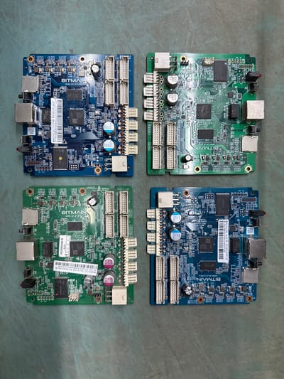 Four electronic circuit boards are laid out on a textured, grey surface. Each circuit board is rectangular and features various electronic components, connectors, and printed text.

The top-left board is blue and has "BITMAIN" printed in white lettering. It includes an RJ45 connector, several smaller connectors, and numerous chips and resistors. A white label with a barcode and text is visible.

The top-right board is green and also has "BITMAIN" printed on it. It features two large black chips, smaller components, and several connectors.

The bottom-left board is green and has "BITMAIN" printed on it. Similar to the top-left board, it has an RJ45 connector, smaller connectors, and a white label with a barcode and text. Two capacitors are prominently visible near the top edge of the board.

The bottom-right board is blue and has "BITMAIN" printed on it. It features two large black chips, smaller components, and several connectors, including an RJ45 connector. A white label with a barcode and text is visible.

The grey surface appears to be a floor or a workbench. The lighting suggests an indoor environment. No people, animals, or any other objects are present in the image. The image is taken from a top-down perspective.

The context of Indiantown, United States of America, is not discernible from the visual information in the image.