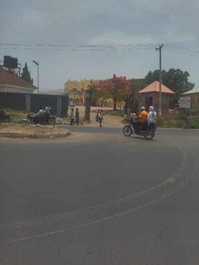 The image depicts a street corner in New Karu, Nigeria. A motorcycle with two people on it is turning onto the street from a smaller side road. The motorcycle is driving on the right side of the road. There are several other people walking on the side of the road. There is a small building with a sign for "Dental Clinic". There is a large tree with red flowers in the background. The sky is overcast. The street is paved and there are tire tracks on the pavement. There is some debris on the side of the road. The location is a residential area with houses and buildings on both sides of the street. There is a metal gate in the background, and the view is from a slight elevation, giving a perspective down onto the street. The street is relatively empty, except for the people and motorcycle.