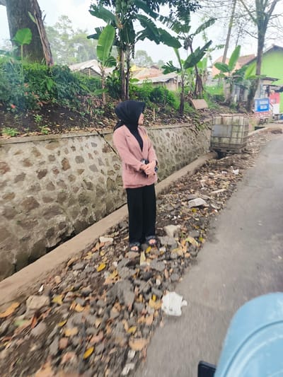 The image shows a woman wearing a black hijab and a pink blazer standing on a paved road in Lembang, Indonesia (latitude: -6.835, longitude: 107.602). The woman is facing away from the camera and is looking to the right. She is standing on a section of the road that has been partially covered in gravel. Behind her is a low, stone wall topped with a concrete curb. There are trees and houses visible behind the wall. The road is surrounded by vegetation and debris. The image is taken from a low angle, giving the impression of a perspective from a vehicle. The image appears to be taken in daylight, with a cloudy sky. There is a small, gray metal box in the background next to the stone wall. There is a light blue object in the foreground of the picture that appears to be a vehicle, possibly a car, but it is partially obscured from view.