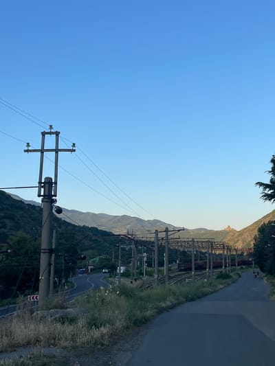 The image depicts a scene in Mtskheta, Georgia, with a clear blue sky dominating the left half. A power line pole with insulators and wires is prominently featured in the foreground. To the right, a winding road curves through a verdant, hilly landscape, with a railway track running parallel to it. In the distance, rugged mountains are visible, their peaks softened by a hazy atmosphere. On one of the distant peaks, a church or monastery with a light-colored dome is perched, a significant landmark. Along the railway, several freight cars are visible, suggesting an active transportation route. A few cars are present on the road, and two figures can be seen walking in the distance on the paved surface. The presence of lush greenery and the overall bright lighting indicate it is daytime, likely late afternoon or early evening due to the warm light hitting the distant mountains. The scene conveys a sense of natural beauty and rural tranquility, with elements of human infrastructure integrated into the landscape. There are no visible text elements in the image.