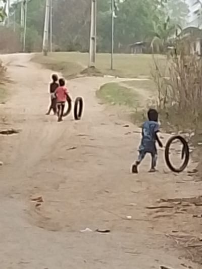 Three children play with tires on a dirt road. Two are together, one wearing a pink shirt. A third child chases a tire in the opposite direction, wearing a blue shirt. The road winds through a village-like area with scattered buildings and utility poles. Vegetation lines the sides of the road. The scene appears to be rural.