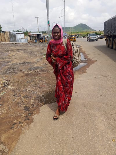 A woman in a red dress and pink hijab stands on a dirt road next to a paved road, wearing a backpack. The background features a street market with various structures, some made of corrugated metal and wood. Several auto-rickshaws are parked, and a truck is driving on the paved road. A distant green mountain is visible under a cloudy sky. The scene appears to be in a rural or semi-urban area. The woman looks directly at the camera, her expression neutral. The ground is a mix of dirt, debris, and puddles, indicating recent rain or poor drainage. The overall impression is a snapshot of everyday life in a Nigerian community, with elements of commerce and transportation. The time of day appears to be daytime, with the sun visible through the clouds.