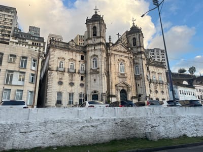 The image displays an urban scene in Salvador, Brazil, centered on the Basílica Santuário Nossa Senhora da Conceição da Praia. This prominent Baroque church, constructed from light-colored, aged stone, features a symmetrical facade with a large arched main entrance, multiple arched windows, and ornate balustraded balconies on its upper levels. Two bell towers, each topped with a dome and a cross, flank the central structure. Weathering and dark streaking are visible on the stone surfaces.
To the left of the church, a multi-story building with a light-colored facade and numerous rectangular windows stands adjacent, partially obscuring other modern high-rise buildings in the background. To the right of the church, a lower white building is visible, followed by an elevated stone retaining wall with some small trees and shrubs on top. A tall, black lamppost equipped with multiple light fixtures and a vertical banner stands on the far right, partially obscuring the view.
In the immediate foreground, a rough, textured white concrete or stone retaining wall runs horizontally across the bottom of the frame, separating a narrow strip of green grass from a paved street. Several passenger vehicles are parked along this street in front of the church, including a white Chevrolet SUV on the left, a light-colored sedan, and multiple darker-colored vehicles further down. The sky is partially cloudy, showing areas of blue between white and grey clouds, and small birds are visible in flight near the church towers. No people are clearly discernible.