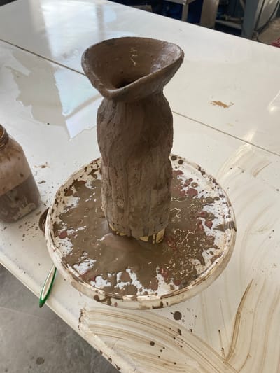 The image shows a clay sculpture on a pottery wheel, likely in a studio setting. The sculpture is rough and appears unfinished, with a wide, open top. The wheel is covered in mud and clay residue. The environment includes a white table covered in splashes and a glass jar. The location is not discernible from the image.
