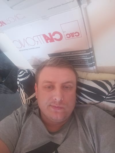 The image is a selfie taken indoors, likely in a residential setting. A man with light brown hair and fair skin is the primary subject, positioned in the lower right quadrant of the frame. He is wearing a grey t-shirt. Behind him, a black and white patterned pillow is visible. In the background, there are stacked boxes with text that appears to be in German. One box has a red and white logo with the letters "CITC". Another box has text that includes "Kuhlfuilu", "Funktionen", and "BTU". There is no indication of the specific location within Litomyšl, Czechia.