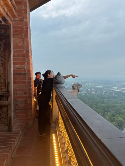 This image captures a view from a vantage point in Tây Hoa Lư Ward, Vietnam, likely from a temple or a high structure. In the foreground, a brick pillar and a carved wooden door are visible on the left. A woman, dressed in black, is leaning on a railing and pointing towards the expansive landscape. Beside her, a man in a black shirt looks towards the camera. The railing is decorated with ornate carvings and illuminated by a strip of yellow lights. Below the railing, a lush green forest stretches out, interspersed with fields and water bodies, with a hazy city in the distance. The sky above is overcast with clouds.